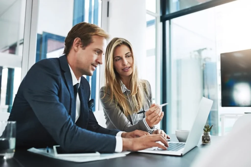Two corporate advisors reviewing financial strategies together on a laptop in a modern glass office, showcasing professional Property Management Consulting Services.