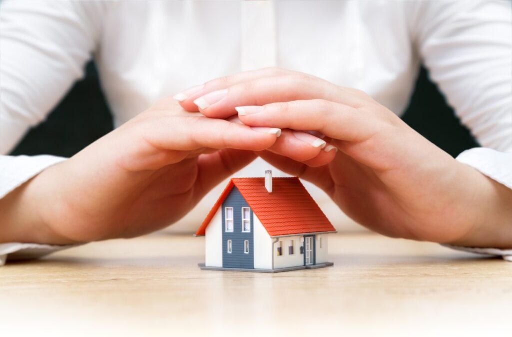 A close-up photograph of a person in a white shirt with hands cupped protectively over a small model house, illustrating the asset protection services offered by a Rental Property Management Company