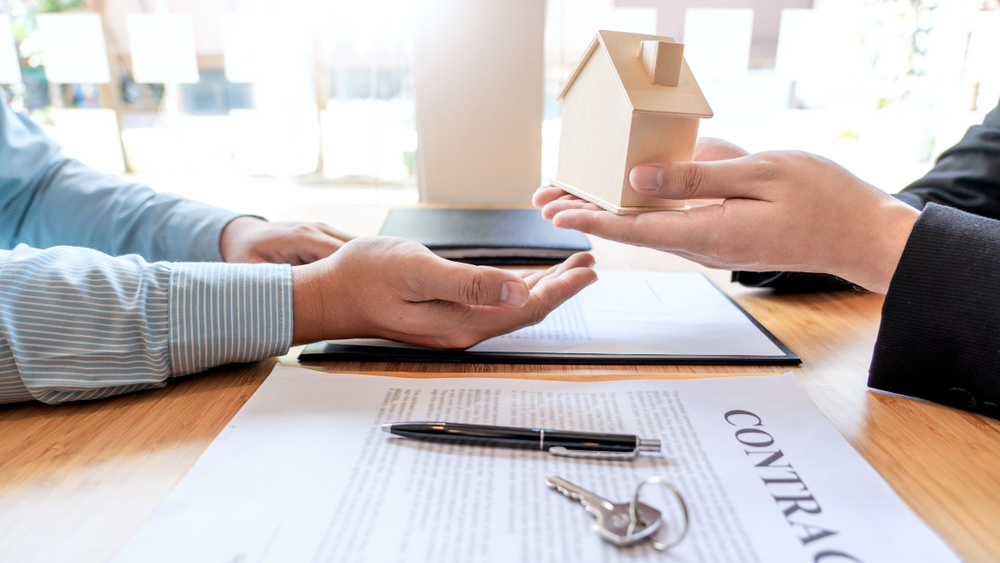 A professional Rental Property Management Company representative in a black suit hands a wooden house model to a client across a desk. In the foreground, a clipboard holds a contract, a pen, and a set of keys, signifying the finalized agreement.